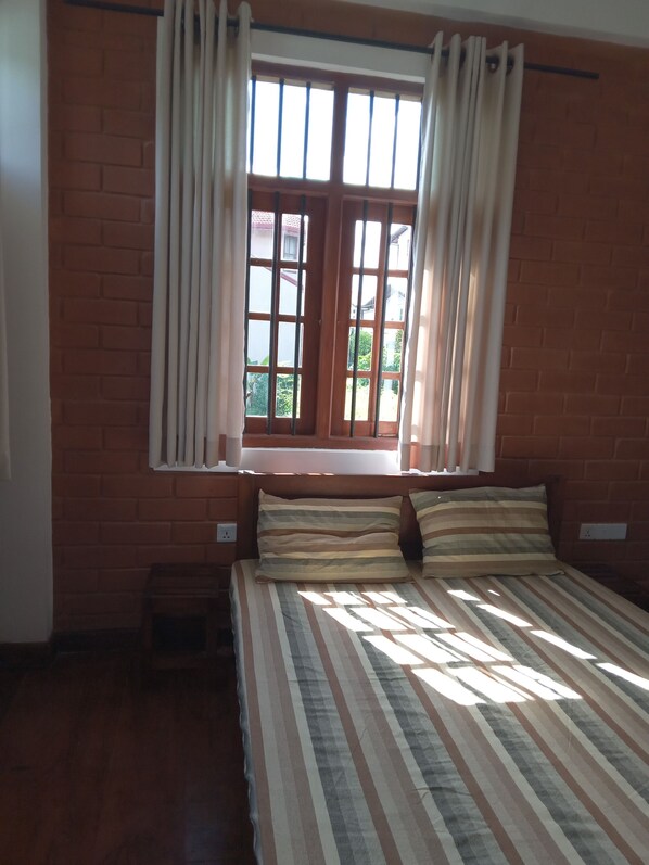 Un dormitorio con pared de ladrillo, una ventana con cortinas, una cama con ropa de cama a rayas y un taburete de madera pequeño.
