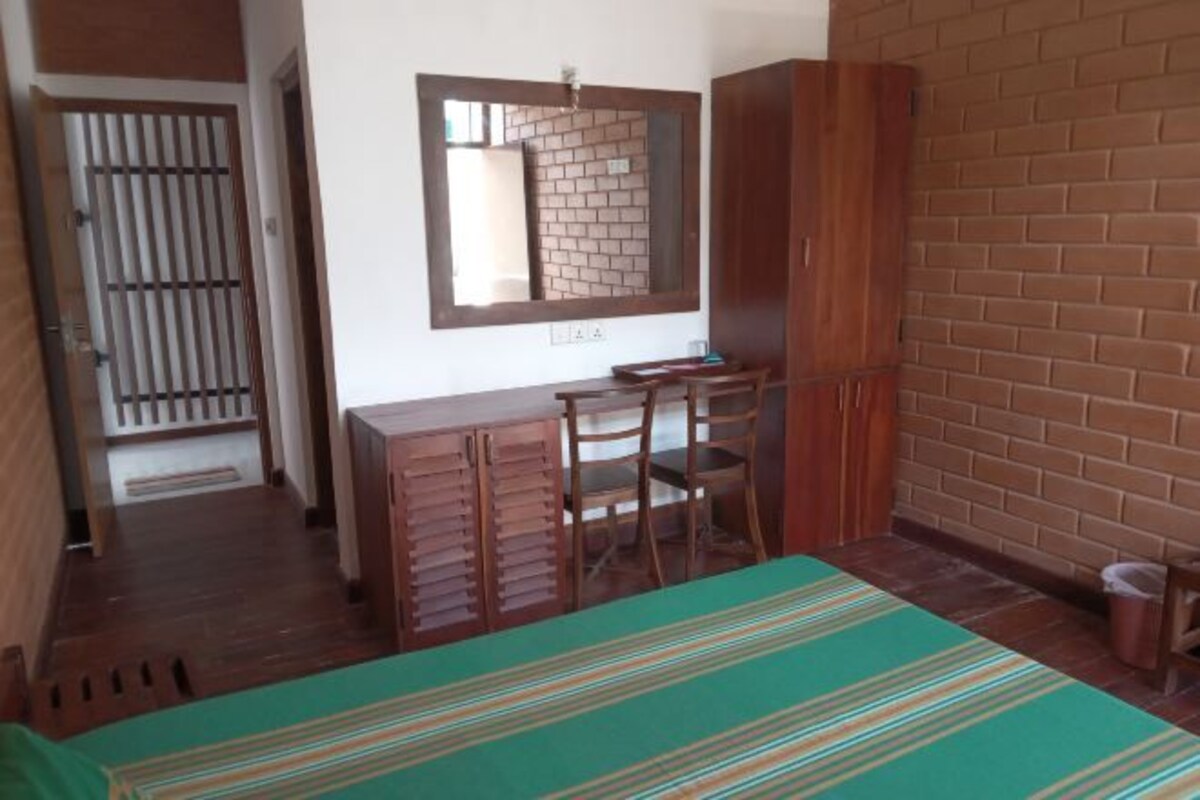 A room with a bed, a desk, and a chair. There is a mirror on the wall, a wardrobe, and a trash bin.