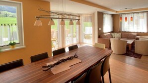 A dining area with a wooden table and chairs, a sofa, and a view of a green landscape through large windows. - Beautiful group accommodation with sauna & ski-in (Itter)