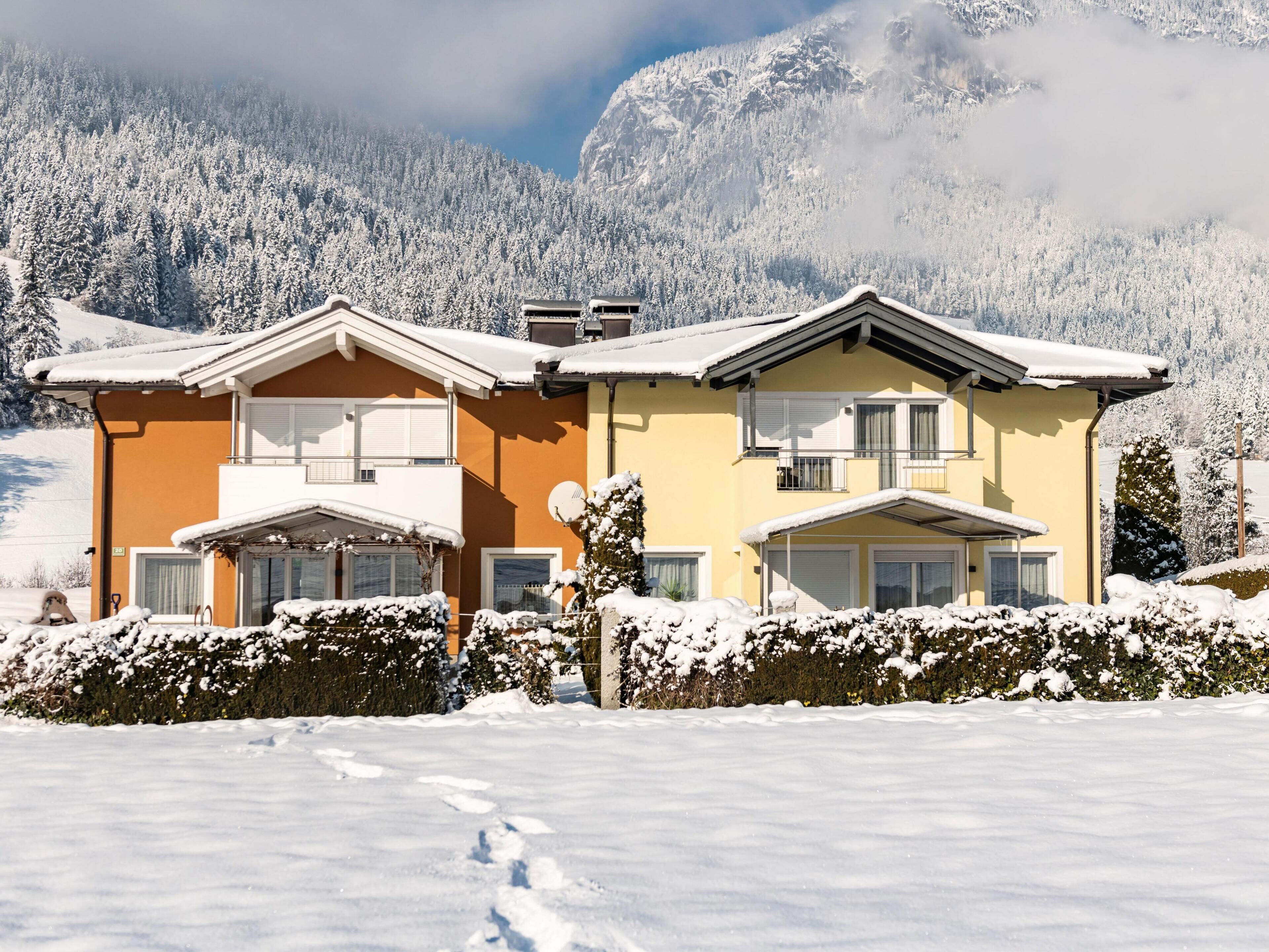 A two-story building with a snow-covered roof, surrounded by snow-covered trees and mountains in the background.