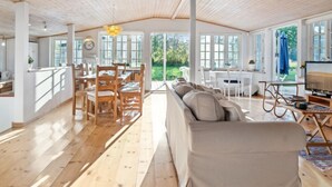 A spacious room with a high ceiling, wooden flooring, a sofa, a dining table with chairs, and a television.