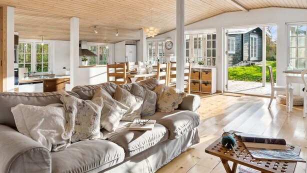 A spacious living room with a large sofa, wooden flooring, and a view of the garden through large windows.