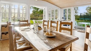 A dining area with a wooden table and chairs, a hanging light fixture, and large windows offering a view of the outdoors.