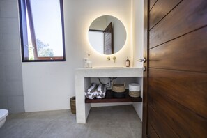 A bathroom with a round mirror, wooden door, and a shelf with baskets. - Beach Villas Canggu 300m from Echo Beach (Canggu)