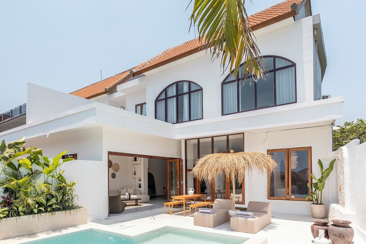 Une maison moderne à deux étages, dotée d’une piscine, d’un espace salon extérieur et d’une grande fenêtre donnant sur la piscine.