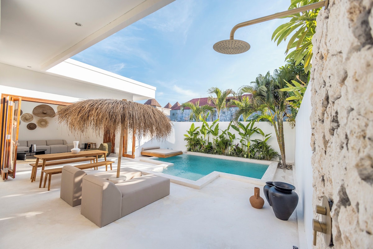 Un espace de vie extérieur moderne, doté d’une piscine, de coins salon et d’un parasol en chaume.