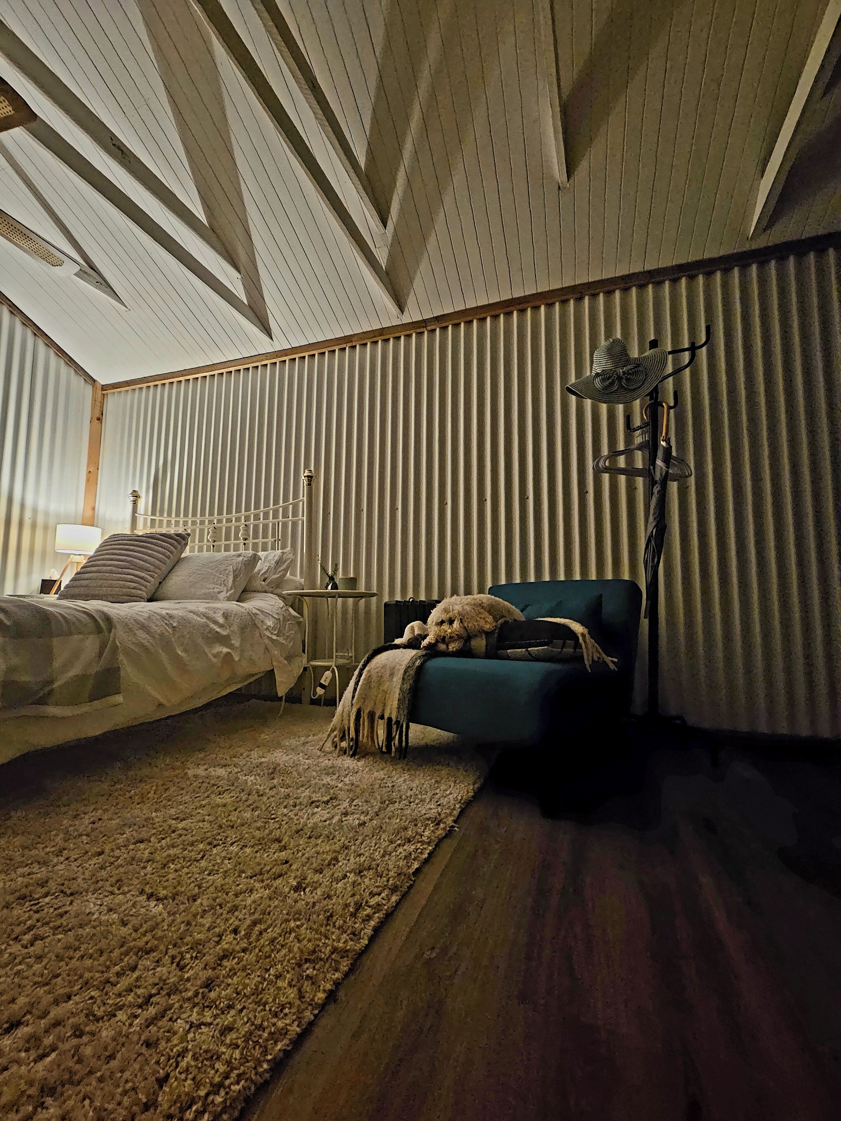 Een slaapkamer met een schuine kap, een bed met hoofdeinde, een blauwe fauteuil, een klein tafeltje en een lamp.