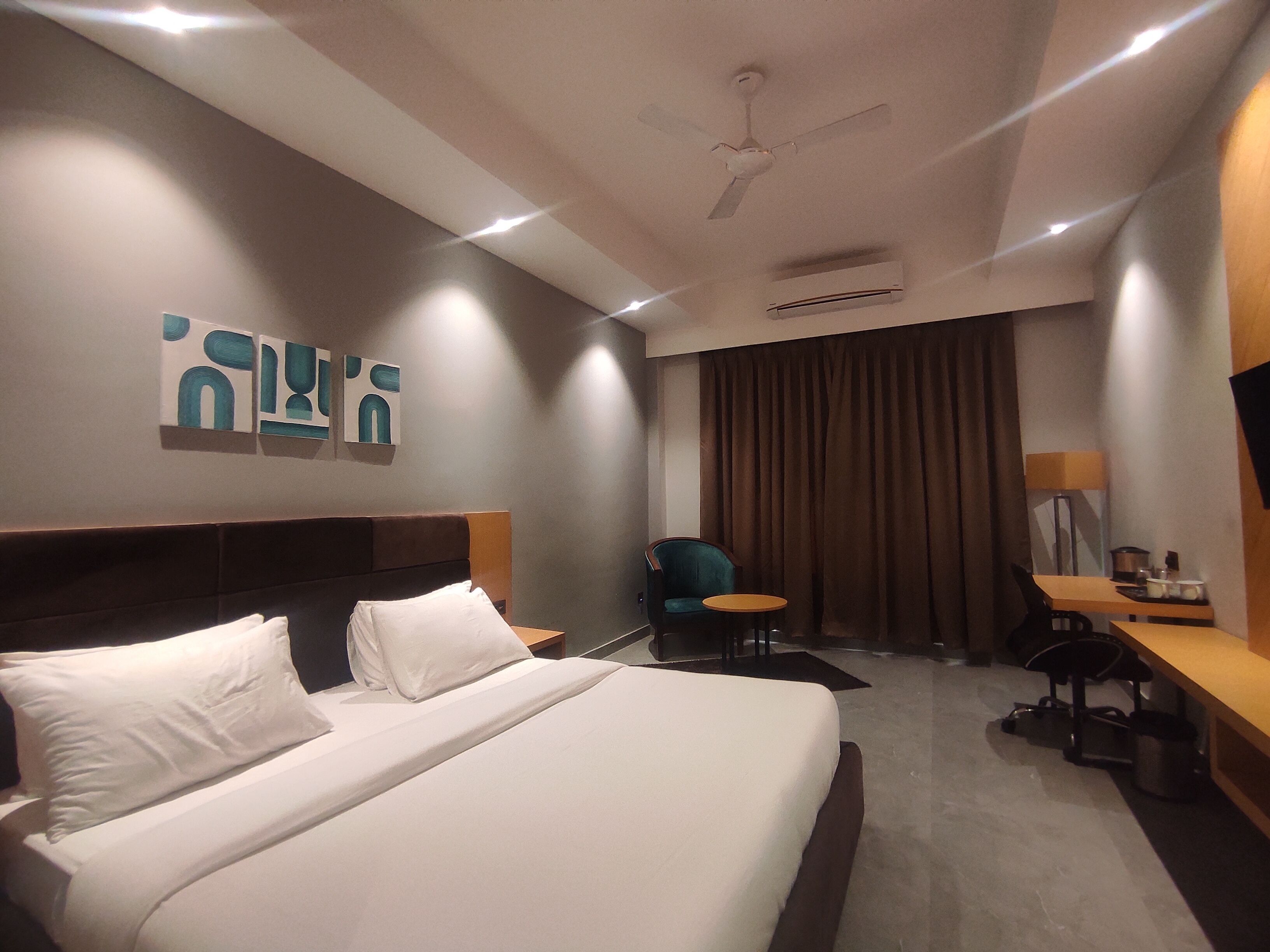 Habitación de hotel con cama, escritorio, silla, ventilador de techo y obras de arte en la pared.