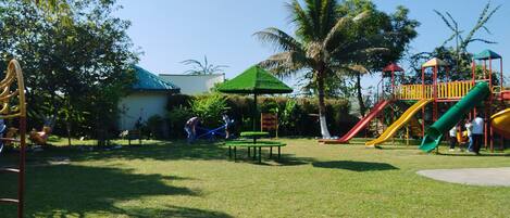 A playground with slides and climbing structures, surrounded by grass and trees.