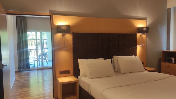 A hotel room with a large bed, two bedside tables, a nightstand, and a view of an outdoor patio. - Padmini Resorts (Tinsukia)