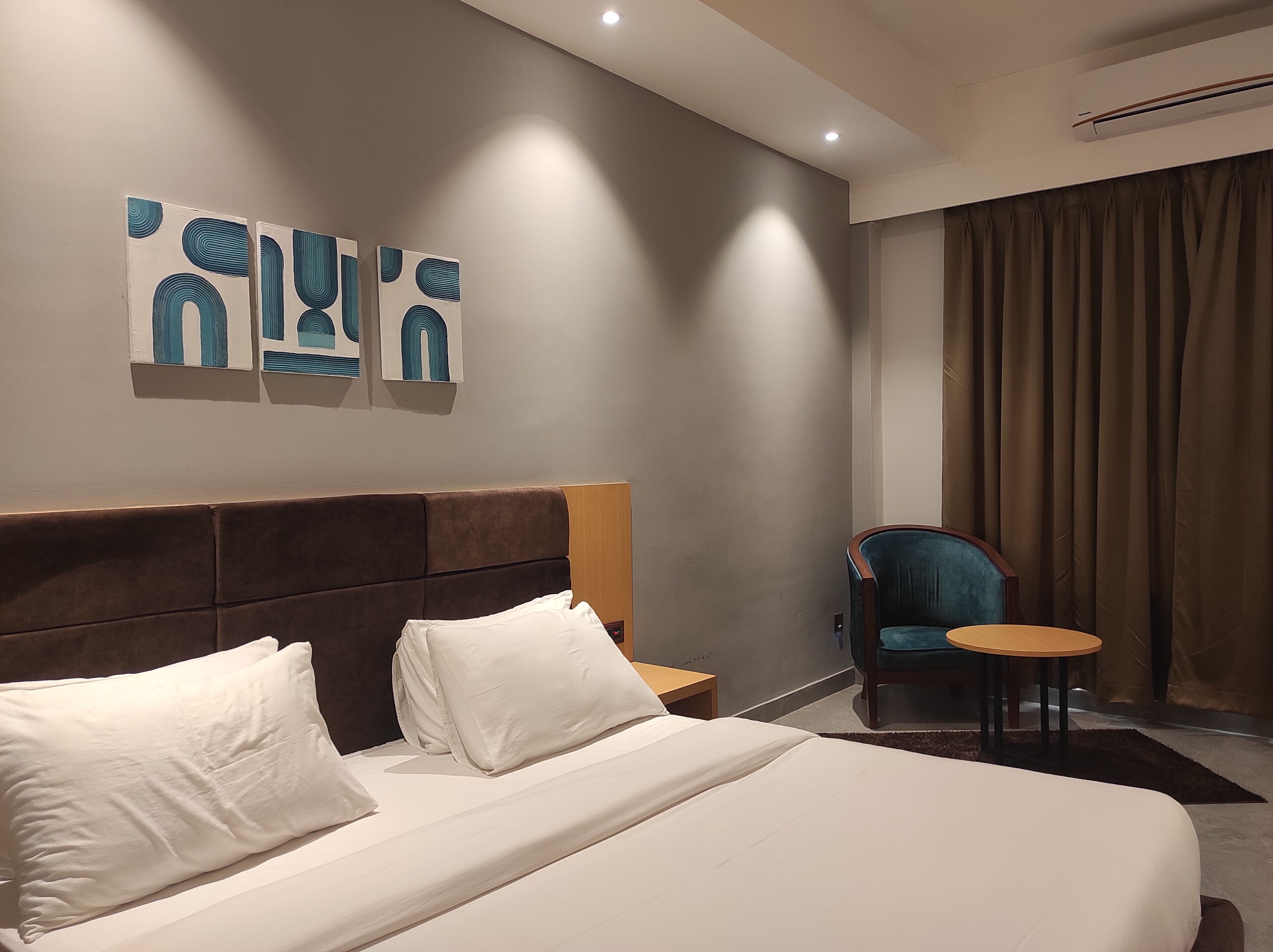 Hotellihuone, jossa on suuri sänky, sängynpääty, pieni pöytä, tuoli ja taideteos seinällä.