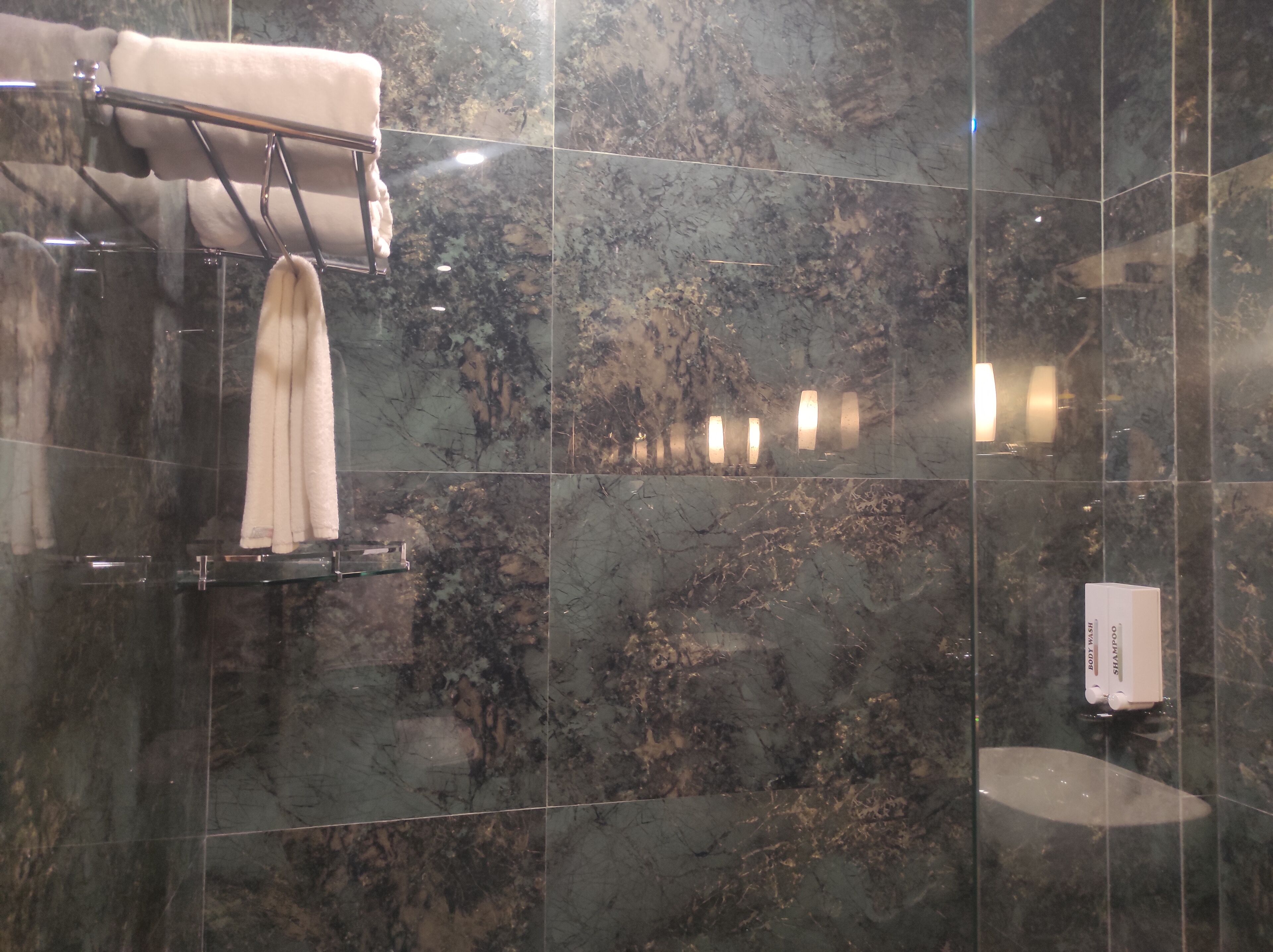 Une salle de bain avec des carreaux de marbre foncé, un porte-serviettes sur lequel est posée une serviette blanche, et une boîte étiquetée « GEL DOUCHE ».