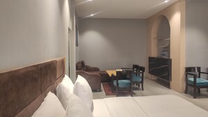 A modern hotel room with a large bed, a sofa, a dining table, and chairs. There is a TV on a stand and a wardrobe in the corner. - Padmini Resorts (Tinsukia)