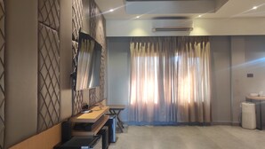 A modern hotel room with a large flat-screen TV mounted on a textured wall, a wooden desk with a computer, a small trash bin, and a large window with sheer curtains. - Padmini Resorts (Tinsukia)