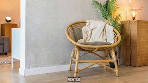 A wicker chair with a cushion, a potted plant, and a wooden cabinet in a room with a concrete wall and a light fixture. - Sandy View - spacious studio flat near the beach (Nieuwpoort)