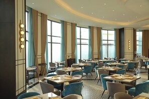A hotel dining area with multiple tables and chairs, large windows with curtains, and a modern ceiling design.