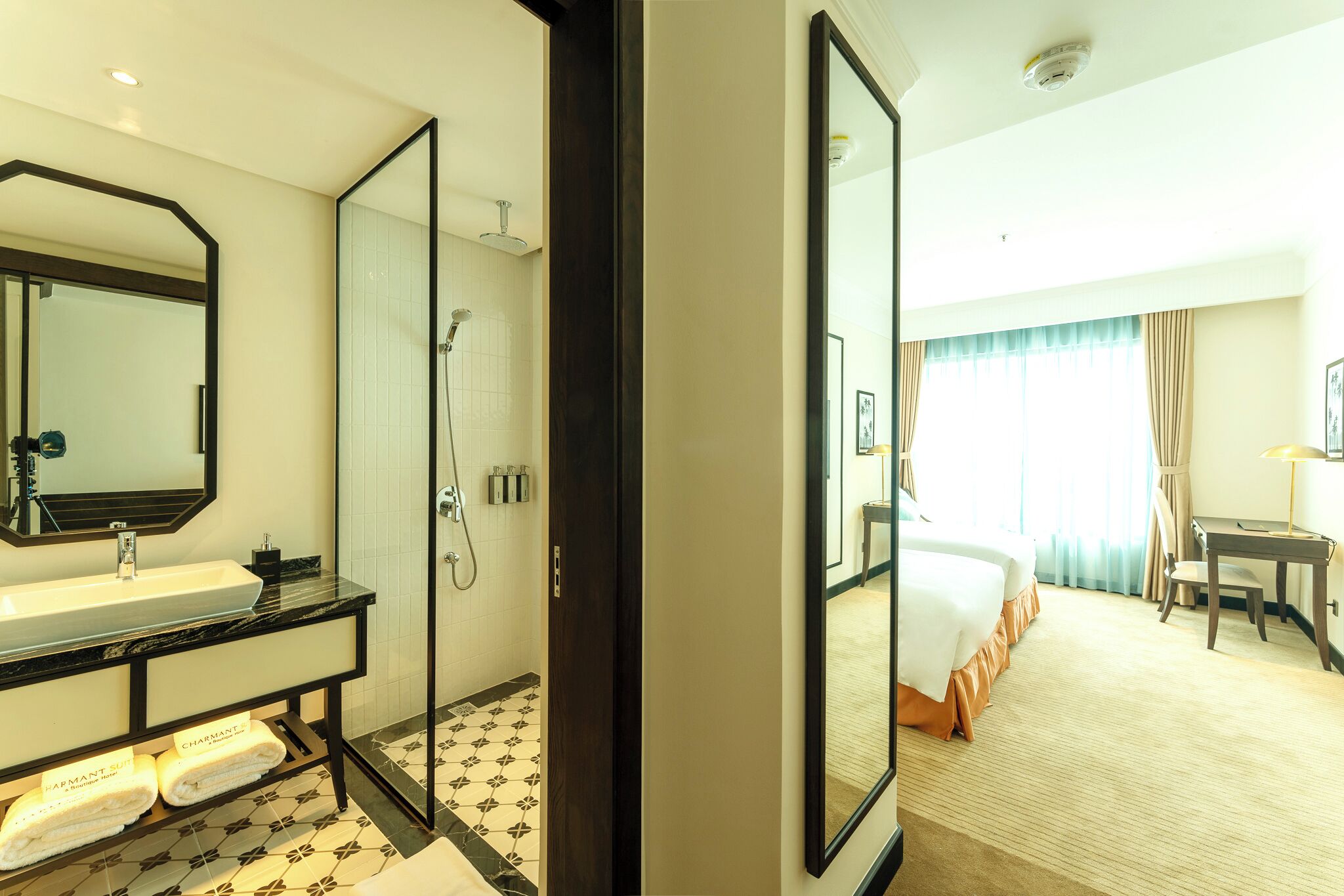 Quarto de hotel com um espelho grande, pia, chuveiro, cama, escrivaninha e uma cadeira.