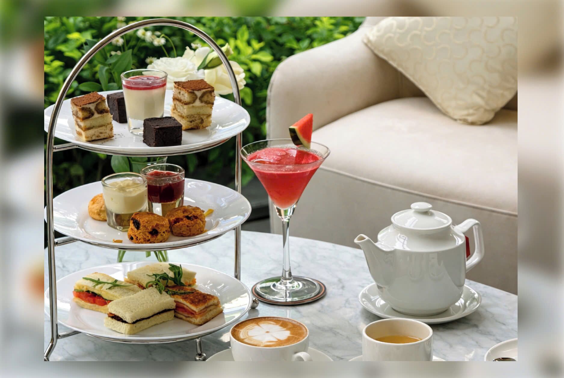 A three-tiered dessert stand with various pastries, a martini glass with a fruit garnish, a teapot, and a cup of coffee on a table.