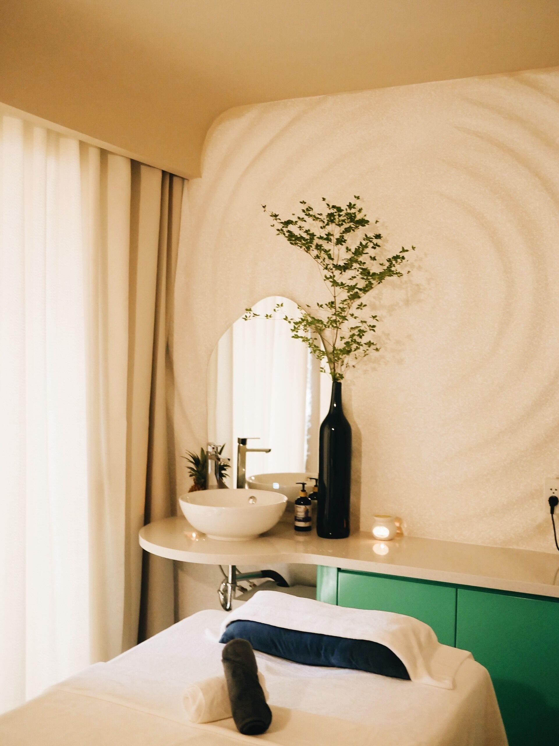 A bathroom with a white sink, a mirror, a green cabinet, and a bed with a white and blue pillow.
