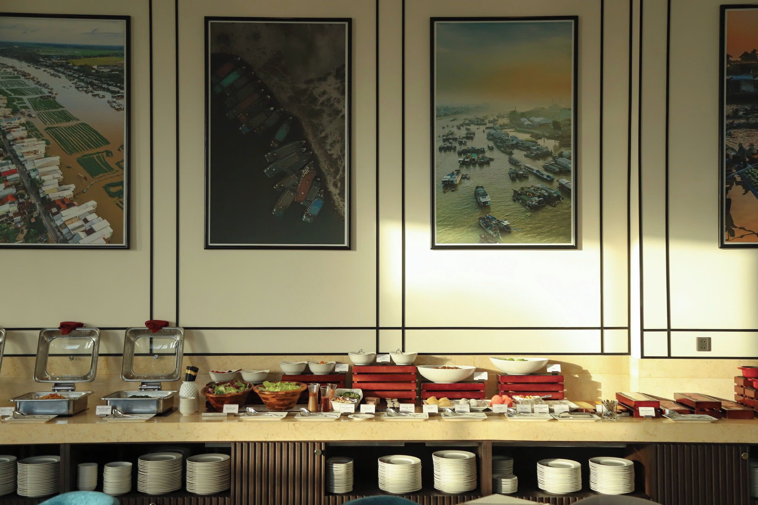 Disposición de buffet con diversos alimentos y recipientes, una barra con platos, y tres grandes cuadros en la pared.