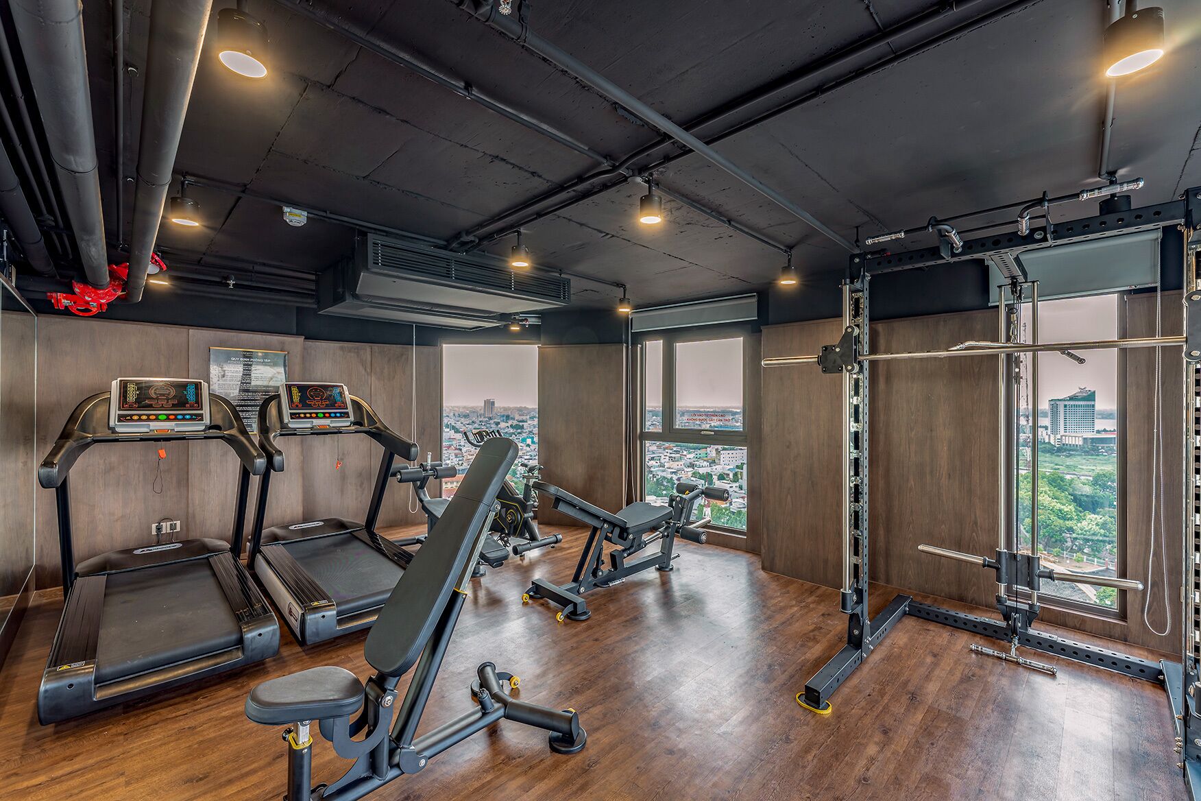 Un gimnasio con cintas de correr, una prensa de banca y una estructura para sentadillas, con amplias ventanas que ofrecen vistas a la ciudad.