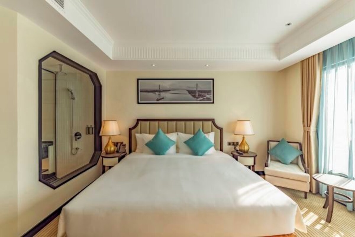 A hotel room with a large bed, two bedside tables with lamps, a chair, a large mirror, and a painting on the wall.