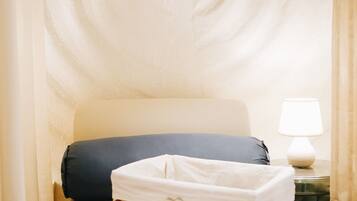 A neatly made bed with a white pillow, a dark blue pillow, and a white towel in a woven basket. A small table with a white lamp is beside the bed.