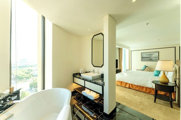 A modern hotel room with a large window, a bathtub, a vanity with a mirror, a bed, and a bedside table with a lamp.