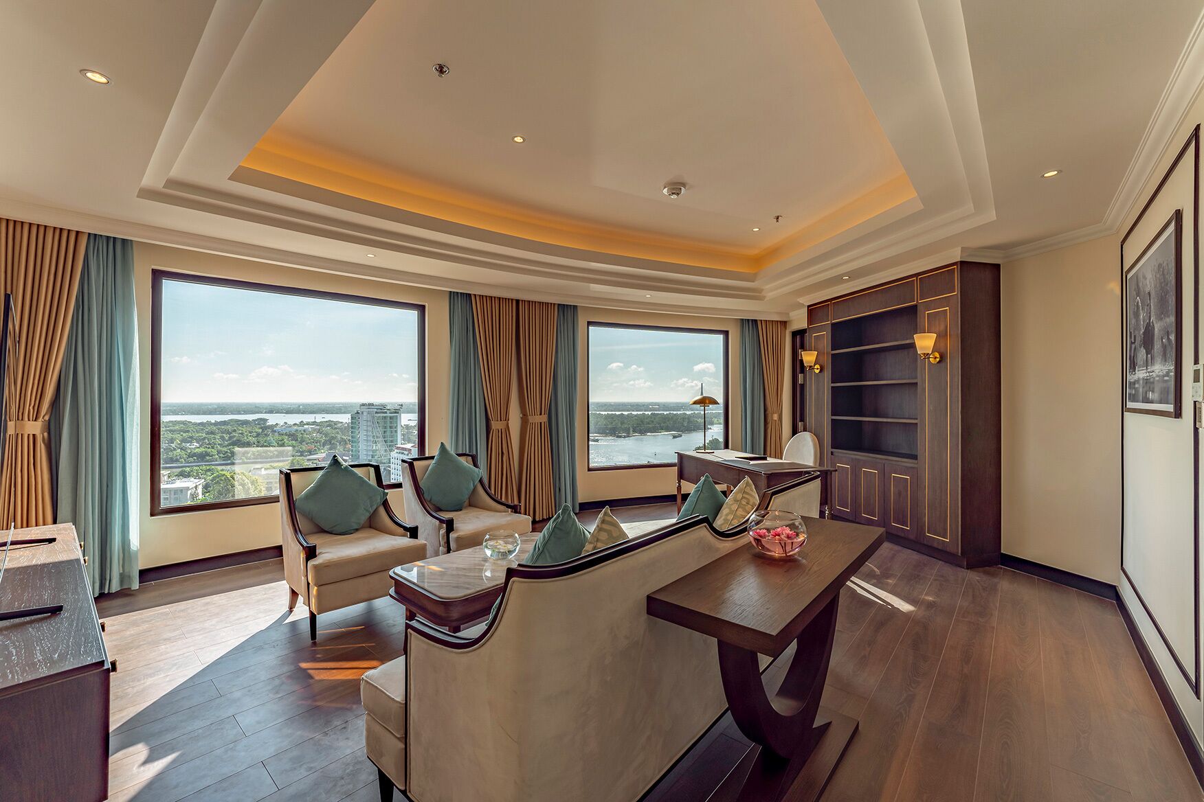 A spacious hotel room with a large window offering a city view, a comfortable seating area with a table, and a bookshelf.