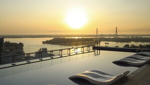 A rooftop infinity pool with lounge chairs, overlooking a cityscape and a river at sunset.