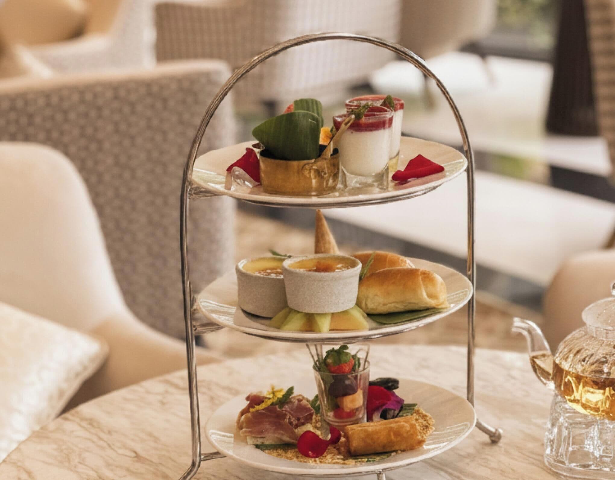 A three-tiered dessert stand with various pastries and desserts, a teapot, and a cup of tea on a marble table in a cozy indoor setting.