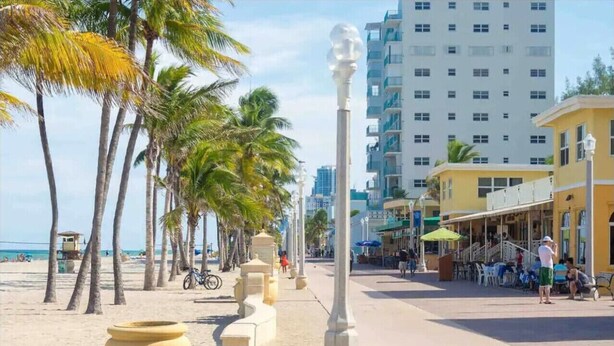 A beachside promenade with palm trees, a café, and a building with balconies. - The Cozy Pelican Studio! Just Steps to the Sand! (Hollywood)