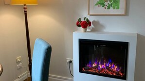 A room with a blue chair, a floor lamp, a fireplace with a fire, and a framed botanical print on the wall. - Beautiful Blue Mountains Garden Flat
 (Katoomba)