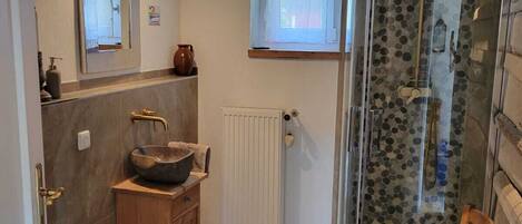 Une salle de bain avec un meuble-lavabo en bois, un grand miroir, une douche dotée d’un pommeau de douche en forme de galet et une petite fenêtre.