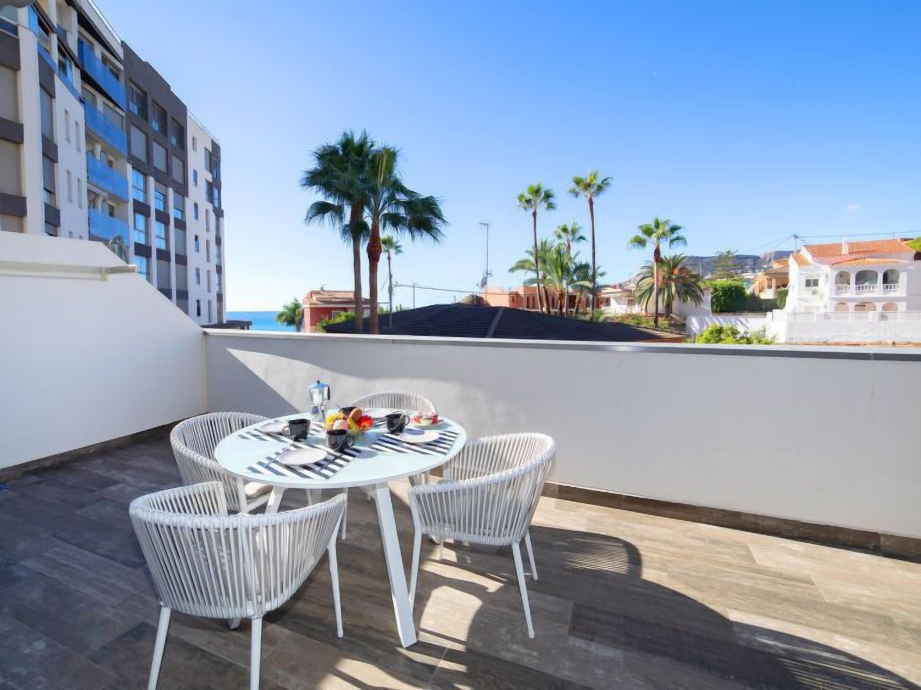 A rooftop terrace with a round table and four white wicker chairs, overlooking a coastal area with palm trees and buildings.