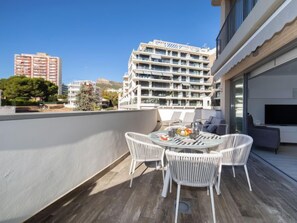 Una terraza en la azotea con una mesa redonda y cuatro sillas de mimbre blanco, con vistas a un paisaje urbano de edificios modernos.