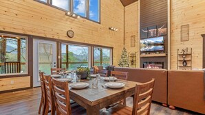 Un interior de madera con una mesa de comedor dispuesta para cuatro personas, una chimenea y un ventanal con vistas a un paisaje pintoresco.