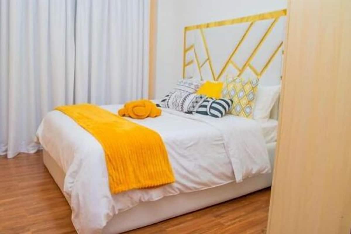 A neatly made bed with a white and yellow bedspread, a yellow throw blanket, and several pillows, including a striped one.