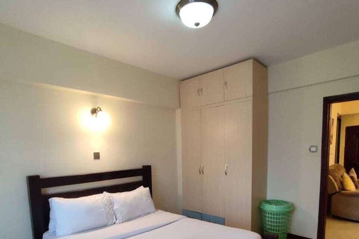 A neatly made bed with white linens and a folded towel, a wooden headboard, a green laundry basket, and a wardrobe with multiple doors.