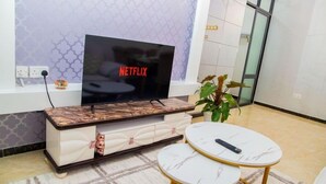Een moderne woonkamer met een flatscreen televisie op een marmeren console, een kleine witte bank, een ronde salontafel en een potplant.