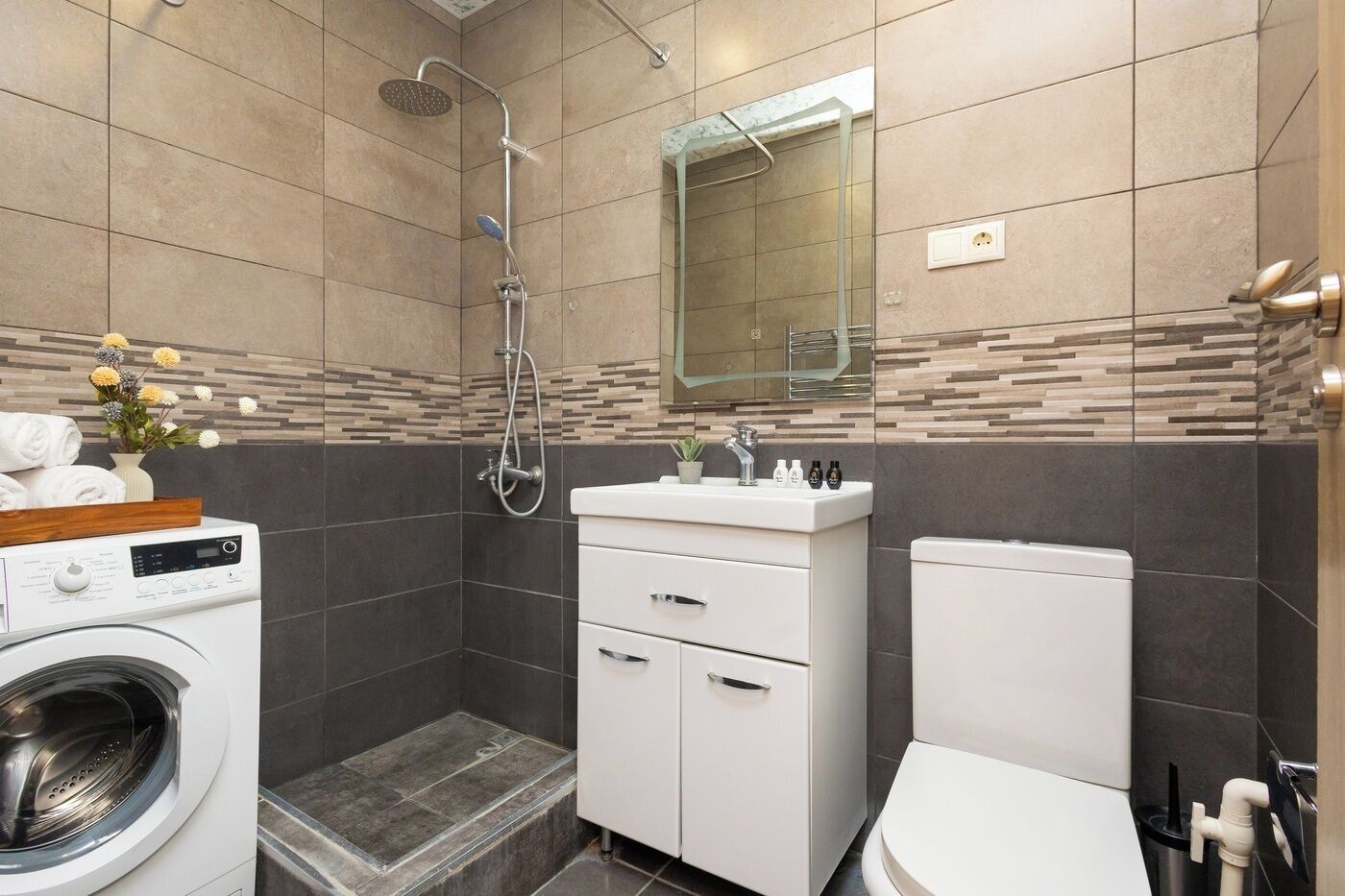 A modern bathroom with a built-in washing machine, a shower, a toilet, and a vanity with a mirror.
