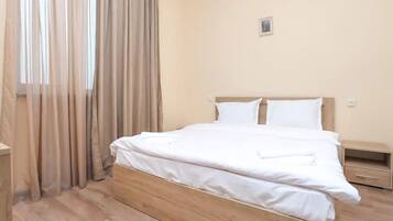 Cama bem arrumada com lençóis brancos num quarto com paredes bege e cortinas.