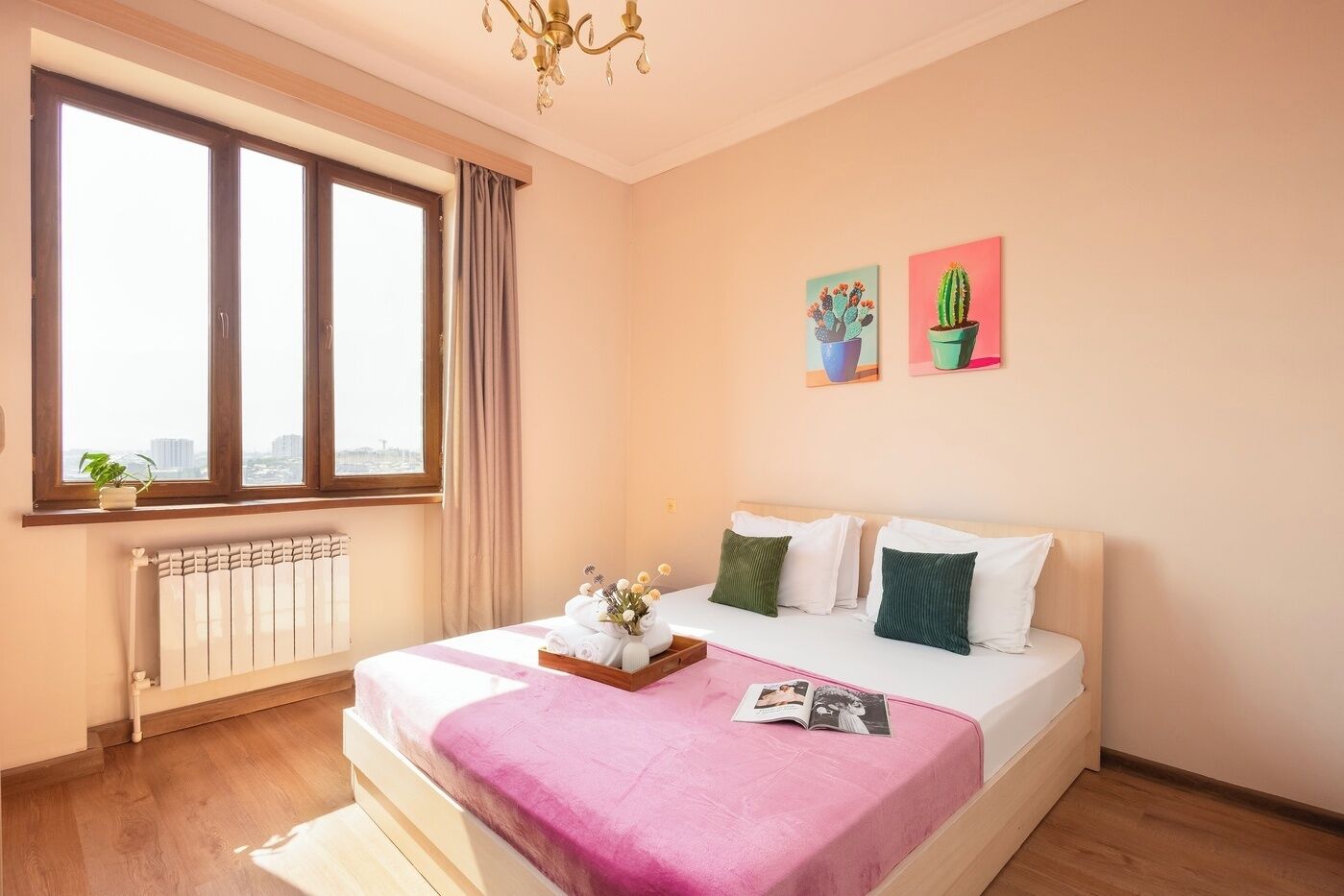 A hotel room with a bed, a window with curtains, a radiator, and two wall-mounted paintings.