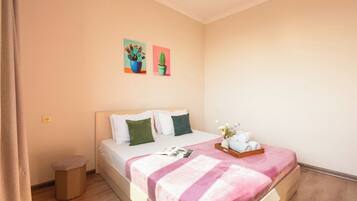 A bedroom with a bed, pillows, a bedside table, a trash bin, and two wall-mounted paintings.