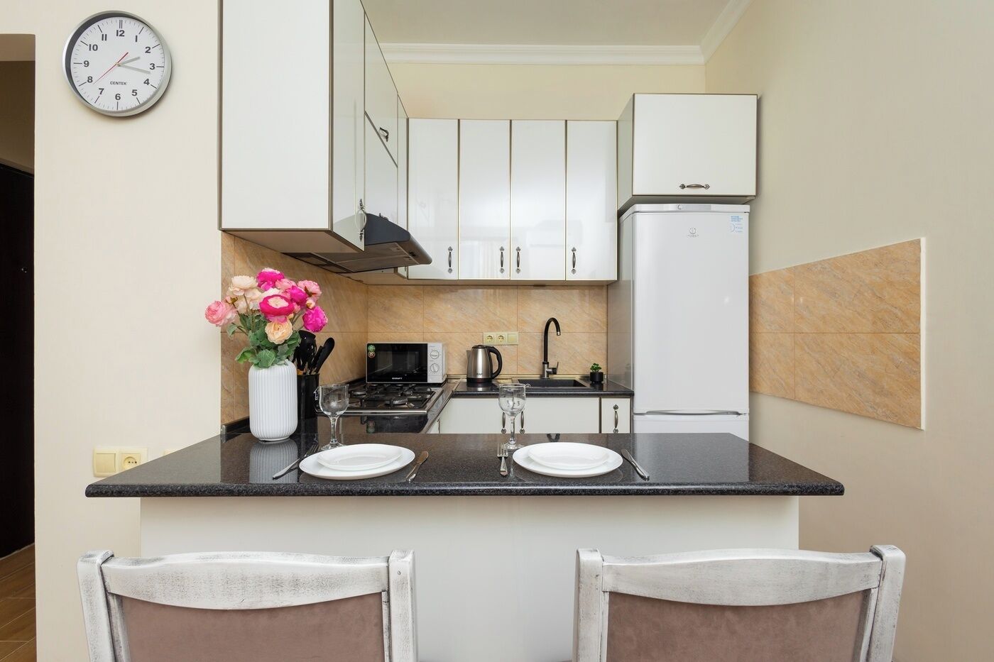 A compact kitchen with white cabinets, a black countertop, a white refrigerator, and a clock on the wall.