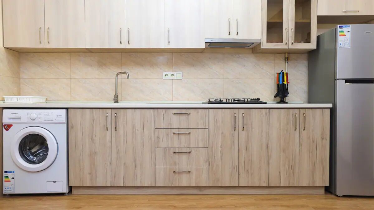 A kitchen with light wood cabinets, a stainless steel refrigerator, a washing machine, and a gas stove.