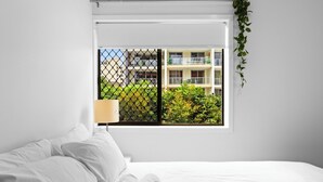A hotel room with a bed, a window with a view of a building and greenery, and a plant on the window sill.