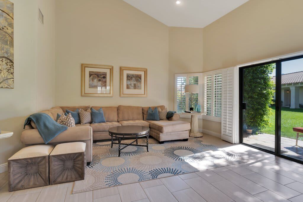 A spacious living room with a sectional sofa, a round coffee table, and a patterned rug. There are two framed artworks on the wall, and the room has a large sliding glass door leading to an outdoor area.