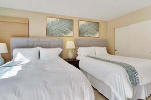 A hotel room with two beds, a nightstand with a lamp, and two framed leaf artworks on the wall.