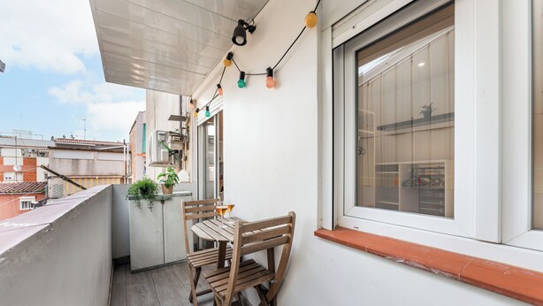 Un balcón con una mesa y sillas de madera, una planta en maceta y una ventana con vistas a edificios.